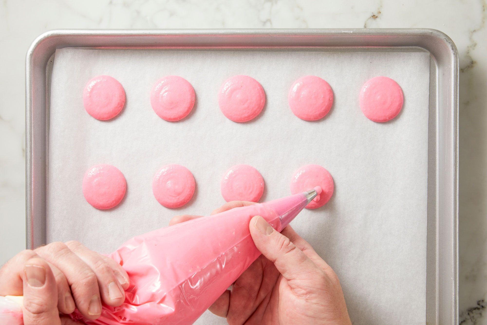 Overhead shot of transfer batter into a pastry bag fitted with a #7 or #10 round tip; Pipe 1-3/8-inch rounds onto a parchment-lined tray about 1 inch apart; Tap tray against counter 2-3 times to remove excess air bubbles; Let macarons rest until no longer wet or sticky to the touch 30-60 minutes; Bake 1 tray at a time until cookies rise about 1/8 inch to form "feet," 14-16 minutes; rotating tray halfway through baking; remove tray and let macarons cool completely; repeat with remaining trays; Once macarons have cooled completely remove from parchment; marble surface;