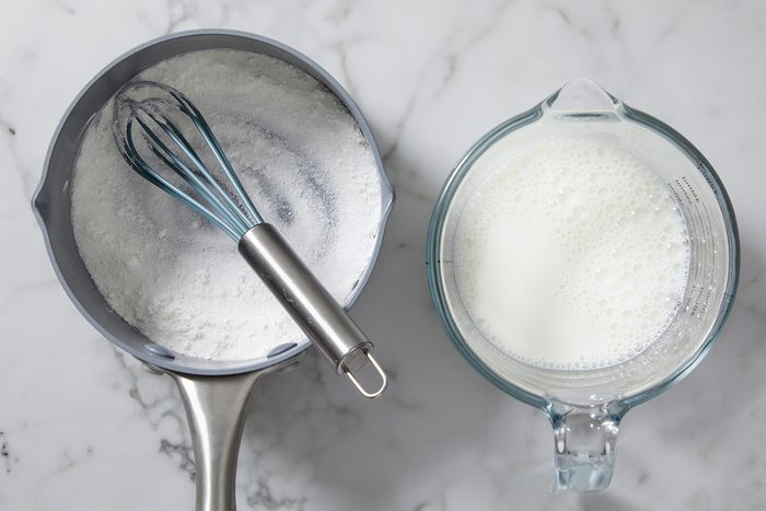 A saucepan with a whisk inside containing a powdery white substance, next to a glass measuring cup filled with milk. Both items are on a marble countertop.