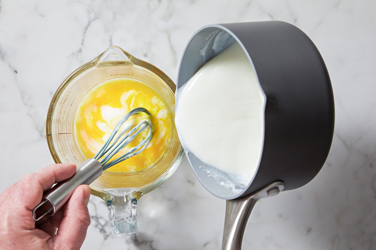 A person pours milk from a saucepan into a glass bowl containing beaten eggs, while holding a whisk. The scene is set on a marble countertop.