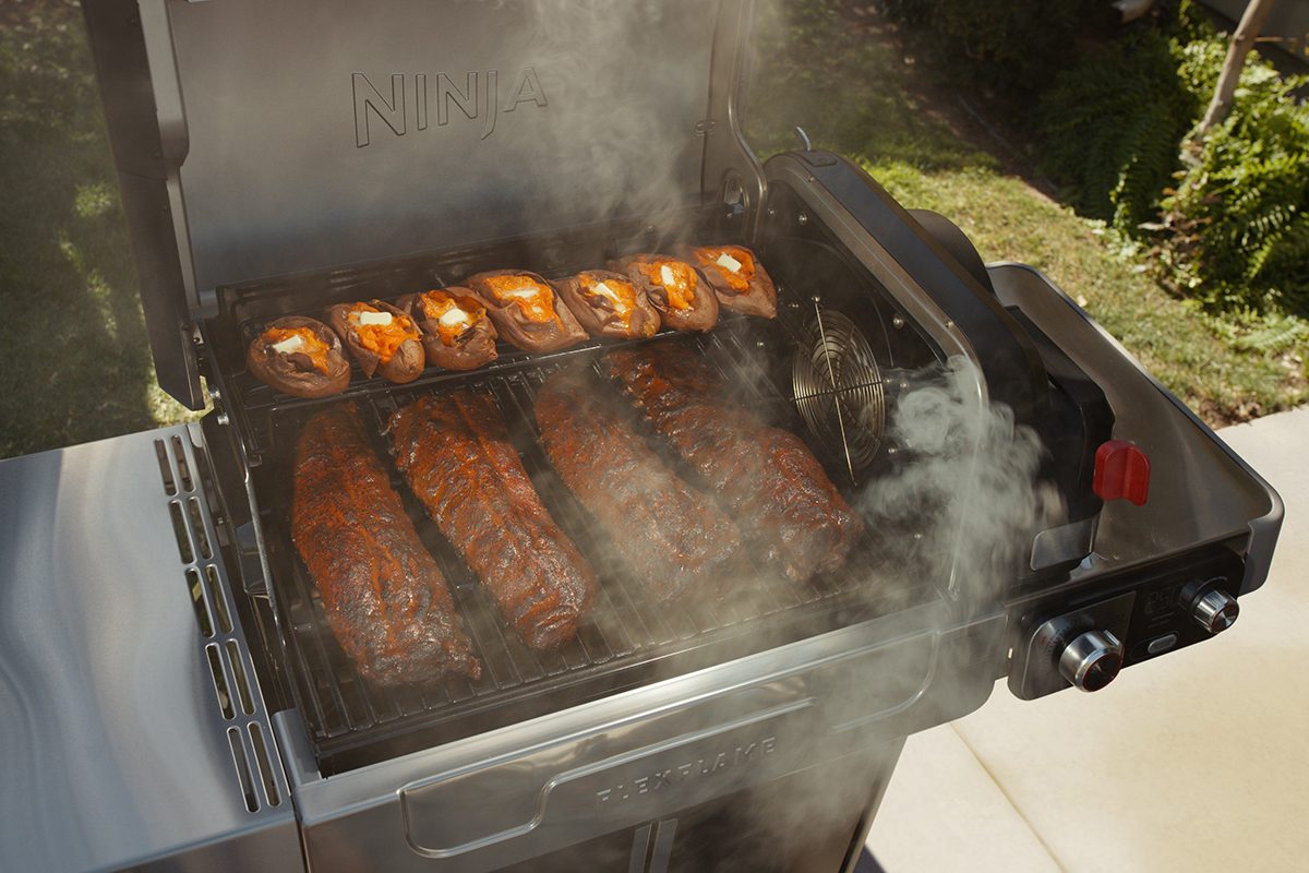 Barbecue grill with smoke rising, cooking several racks of ribs and chicken pieces. The grill has two sections with visible controls on the side, situated outdoors with greenery in the background.