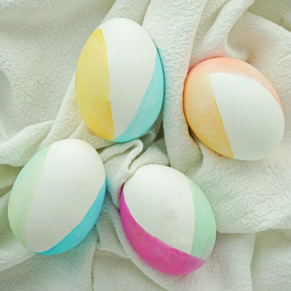 Four pastel-colored eggs resting on a soft, white fabric. Each egg is painted in a two-tone pattern, featuring shades of yellow, blue, orange, green, and pink.