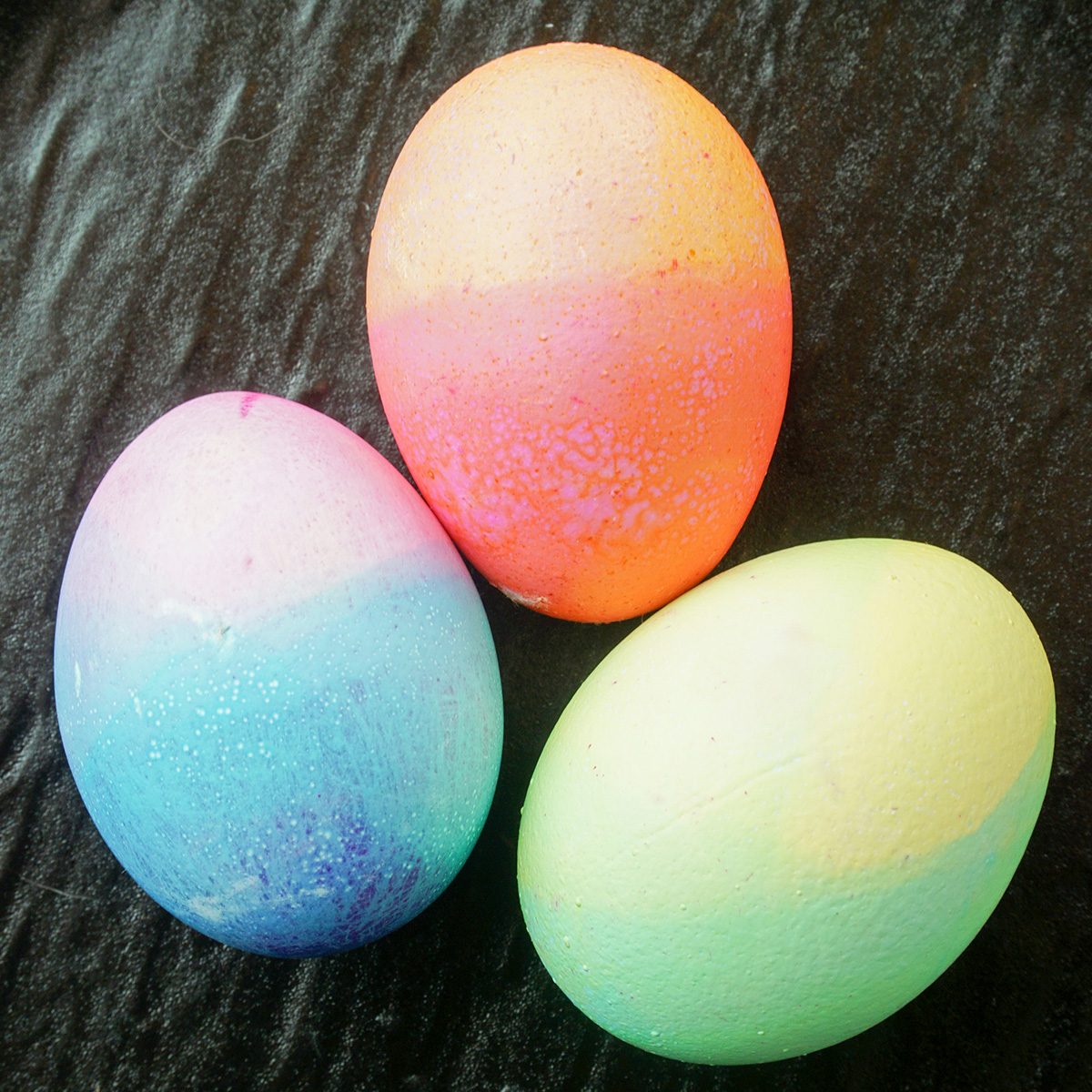 Three vibrantly colored eggs, featuring a gradient of pink, orange, blue, purple, and green hues, rest on a textured dark surface.