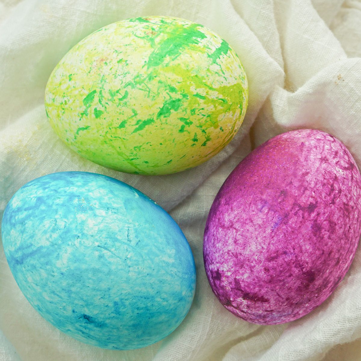 Three colorful eggs with a marbled texture lie on a soft, beige fabric. The eggs are painted in shades of green, blue, and pink, creating a vibrant and artistic display.