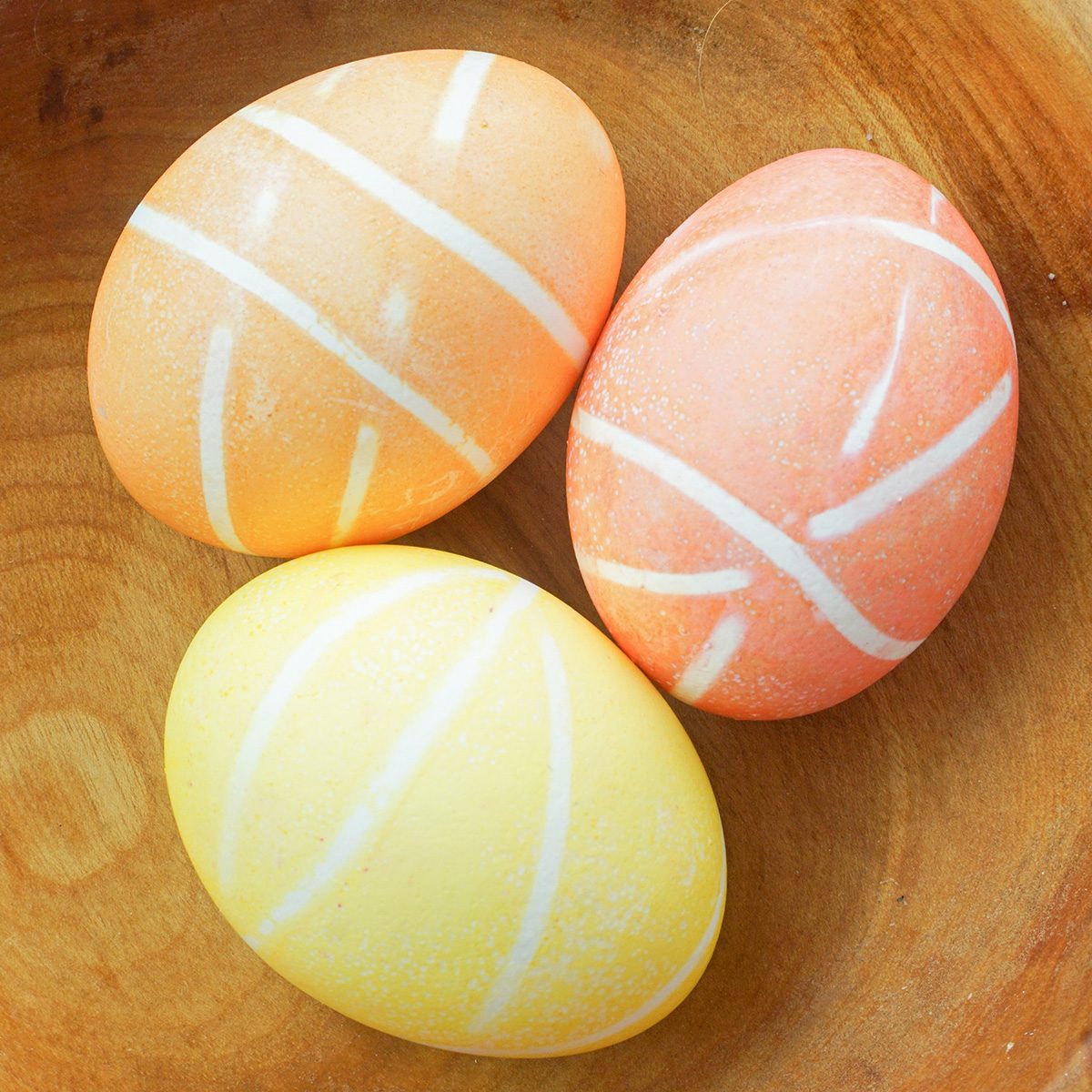 Three pastel-colored eggs with white diagonal stripes are placed in a wooden bowl. The eggs are yellow, peach, and light orange, creating a soft and harmonious color palette.