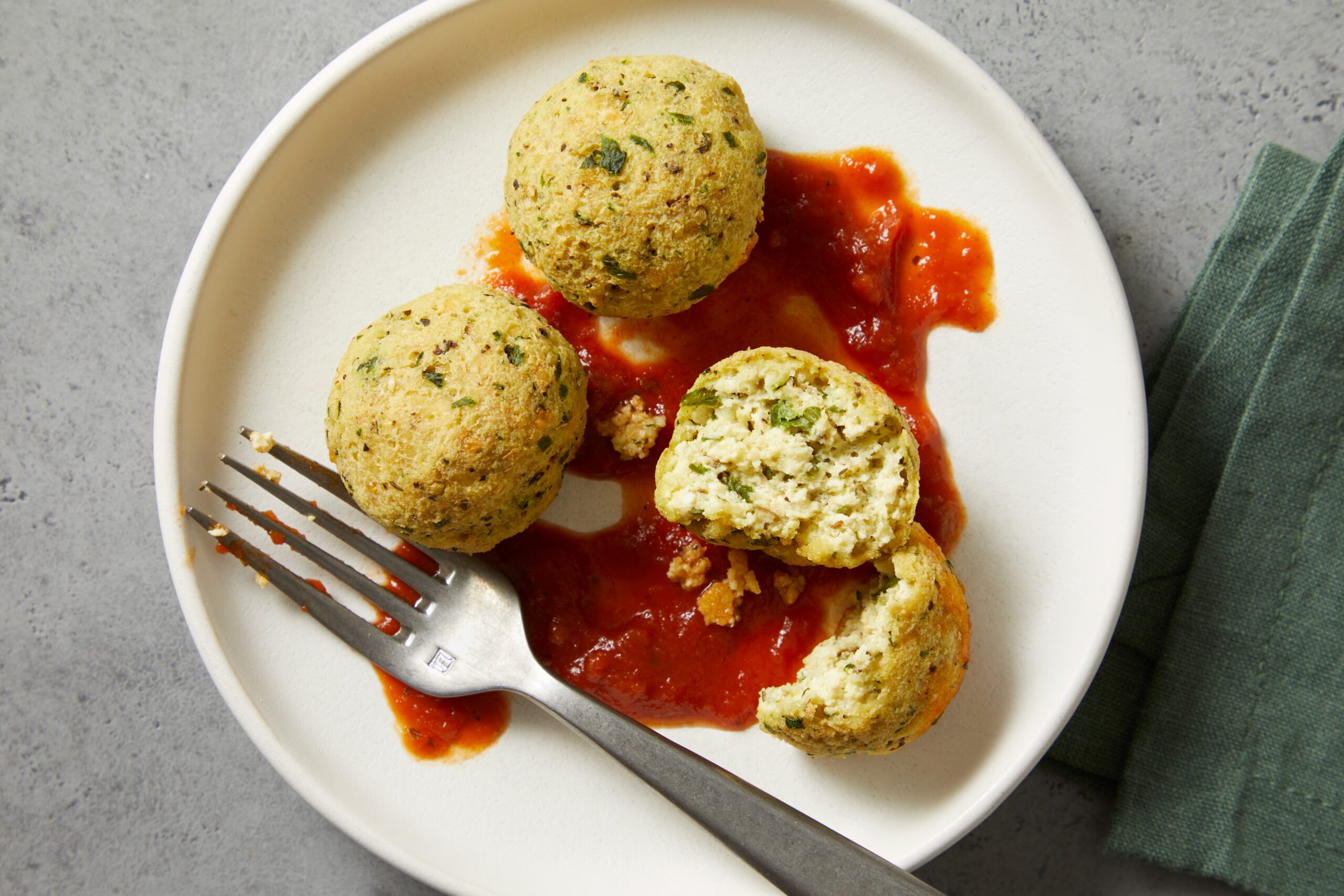A white plate with three broccoli chicken meatballs served on a bed of red sauce. One meatball is partially broken, revealing the inside. A fork rests on the plate, and there's a green cloth beside it. The background is a gray surface.