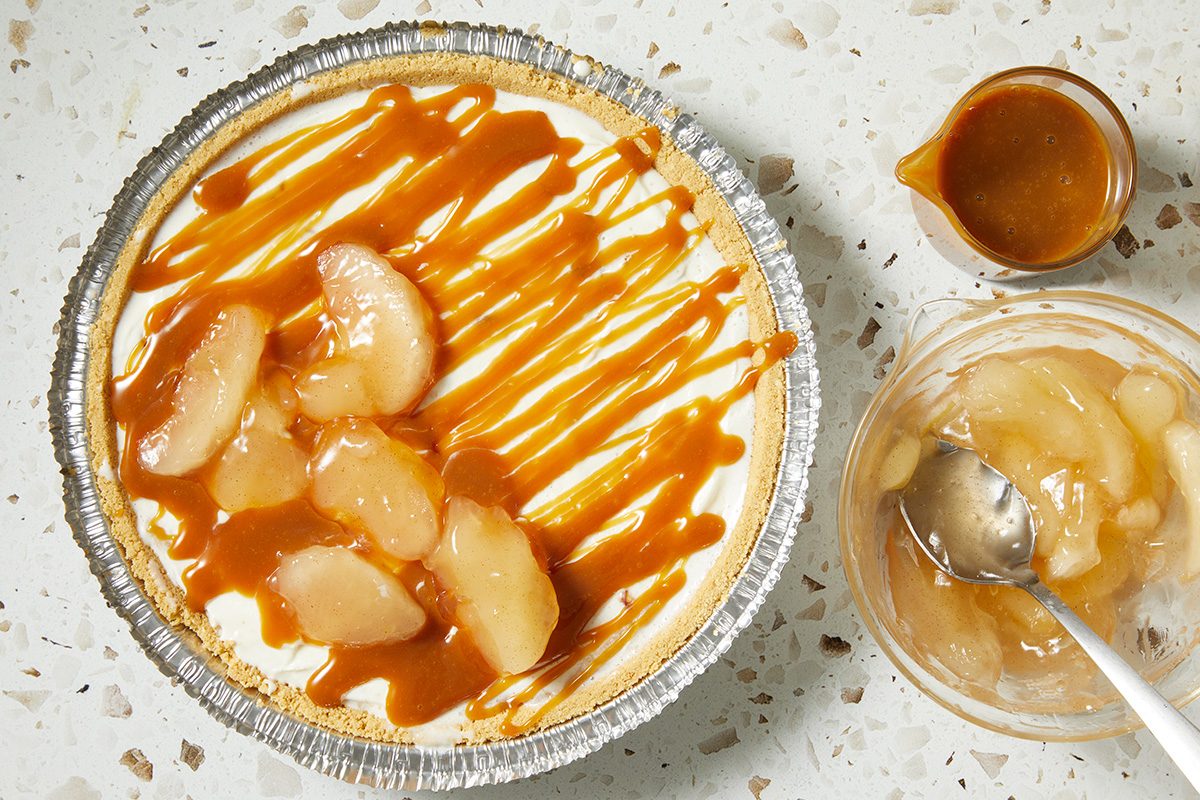 A caramel apple pie in a foil crust, topped with apple slices and caramel drizzle, is pictured next to a bowl of apple filling and a small cup of caramel sauce on a speckled countertop.