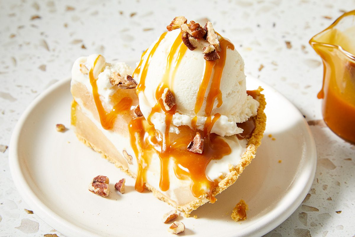 A slice of pie topped with a scoop of vanilla ice cream, chopped nuts, and caramel sauce, served on a white plate with a small container of extra caramel sauce nearby.