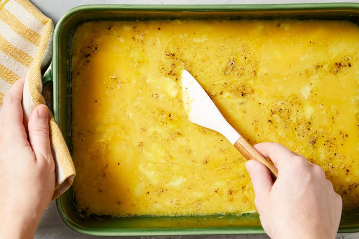 Hands are shown holding a dish and a spatula with cheesy, baked grits inside a green rectangular baking dish.