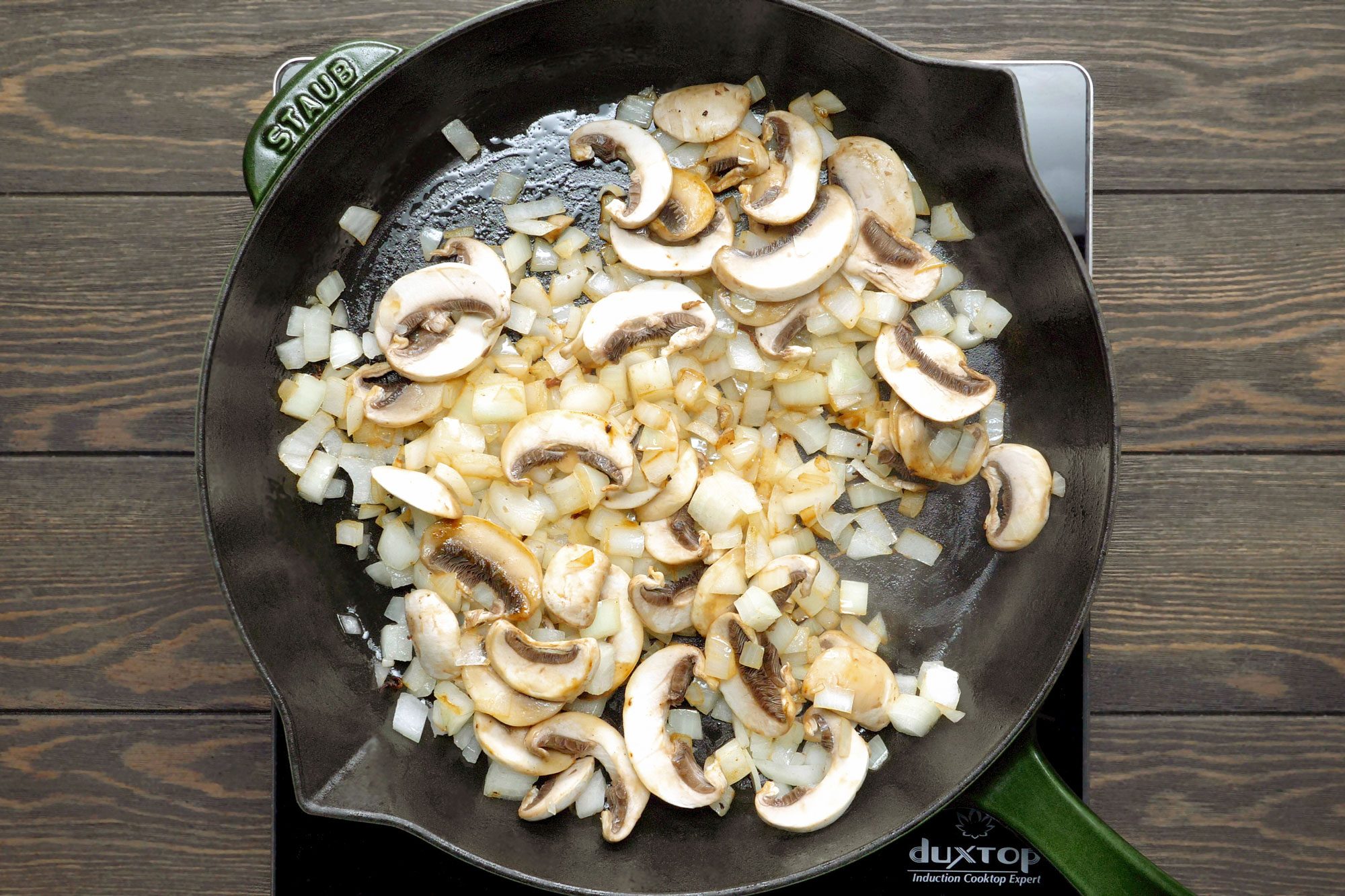overhead shot of a cast iron skillet with a green handle, sitting on a wooden countertop; the skillet is filled with chopped onions and sliced mushrooms that are being sauteed in oil;