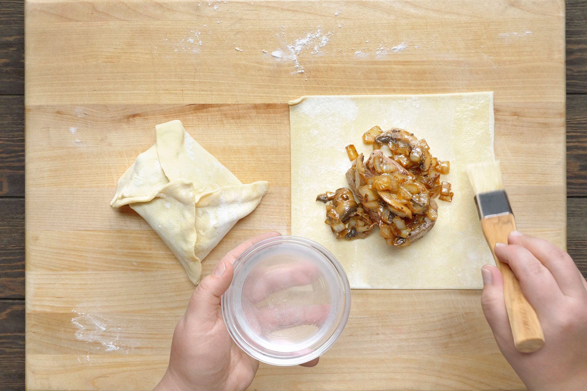 overhead shot of a wooden cutting board with a variety of ingredients on it, likely for a recipe; there is a folded pastry dough, a bowl of water, a piece of meat with sauteed onions and mushrooms and a pastry brush; a hand is holding the bowl of water and a third hand is holding the pastry brush