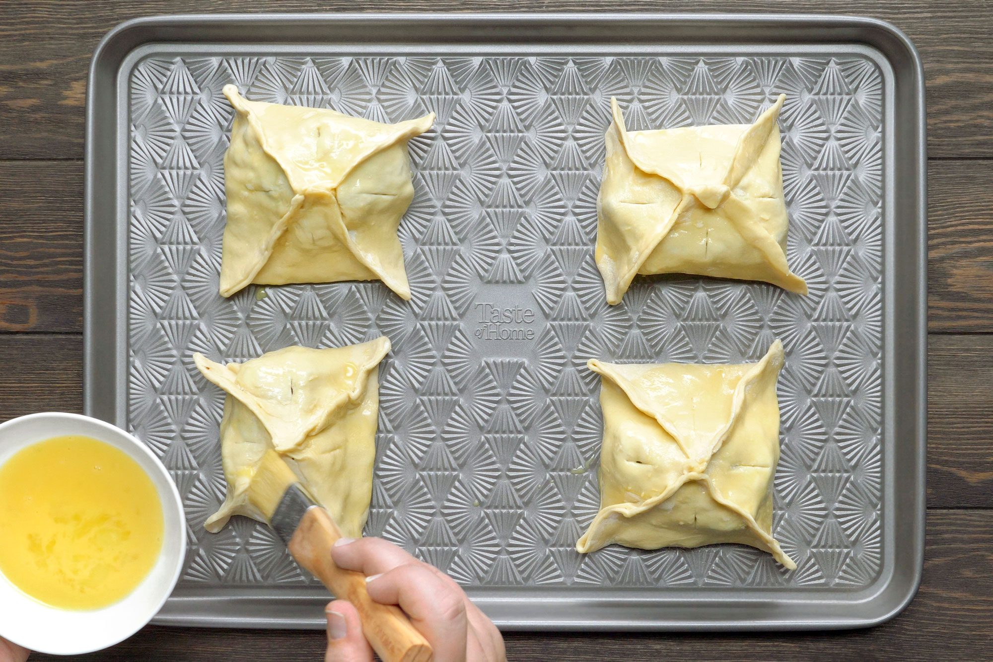 overhead shot of a baking sheet with four pastries on it; the pastries are square-shaped and folded into four points; there is a small white bowl with egg wash in it; a hand is holding a pastry brush and is brushing the egg wash on one of the pastries
