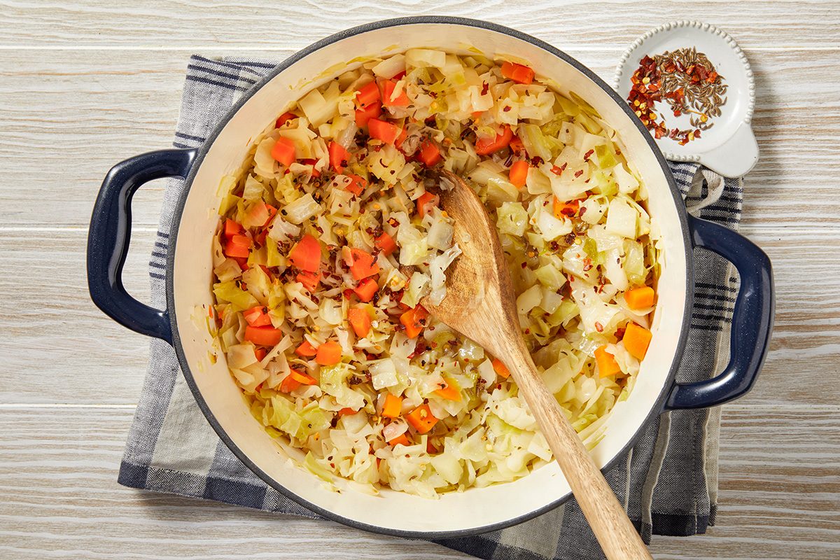 A pot of vegetable soup with chopped carrots, potatoes, and herbs on a table. A wooden spoon rests inside the pot. A small dish with spices is nearby. The pot sits on a dark and white checkered cloth.