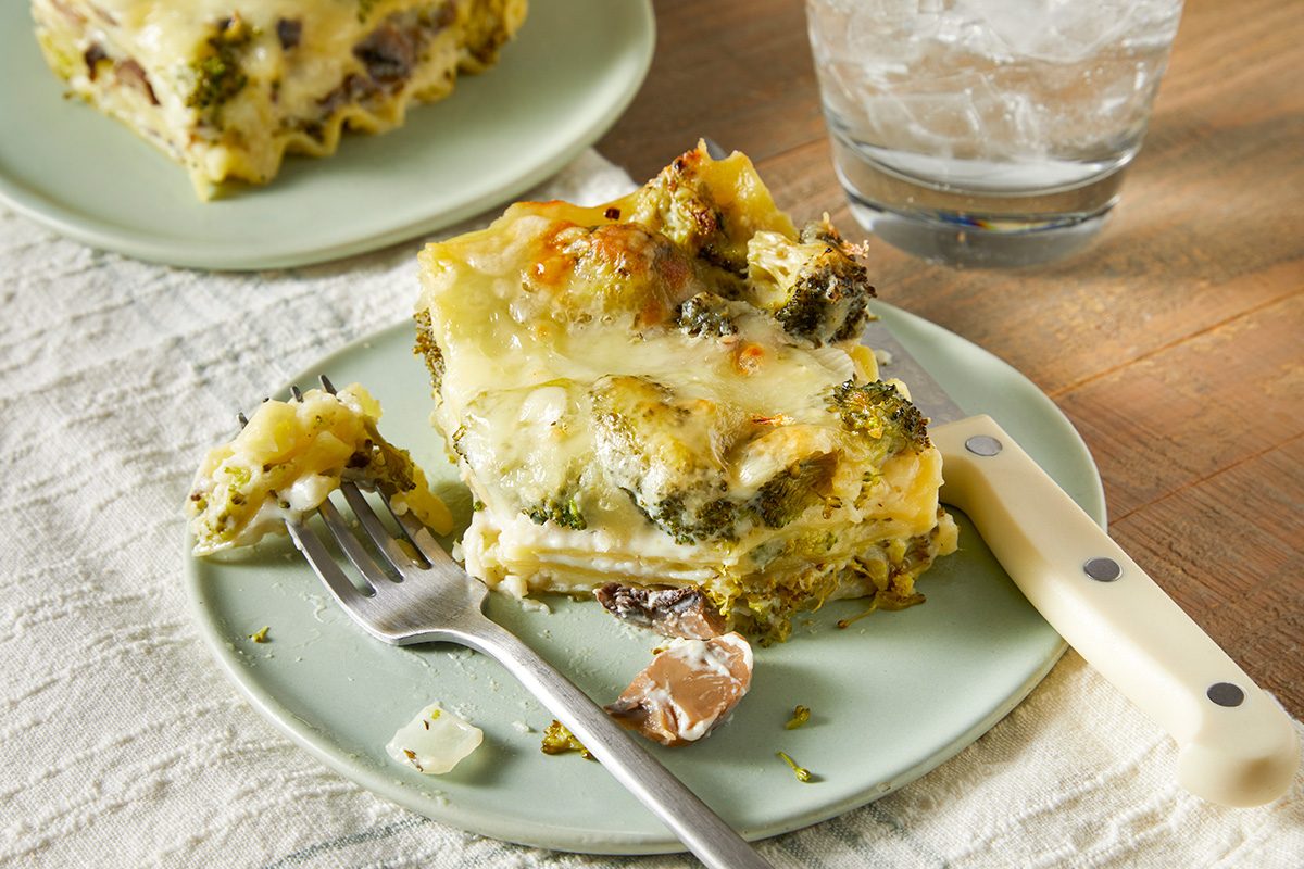 A slice of broccoli and mushroom lasagna on a light green plate with a fork beside it. The lasagna has a golden, cheesy top. Another plate with lasagna is in the background. A glass of iced water sits on the wooden table.
