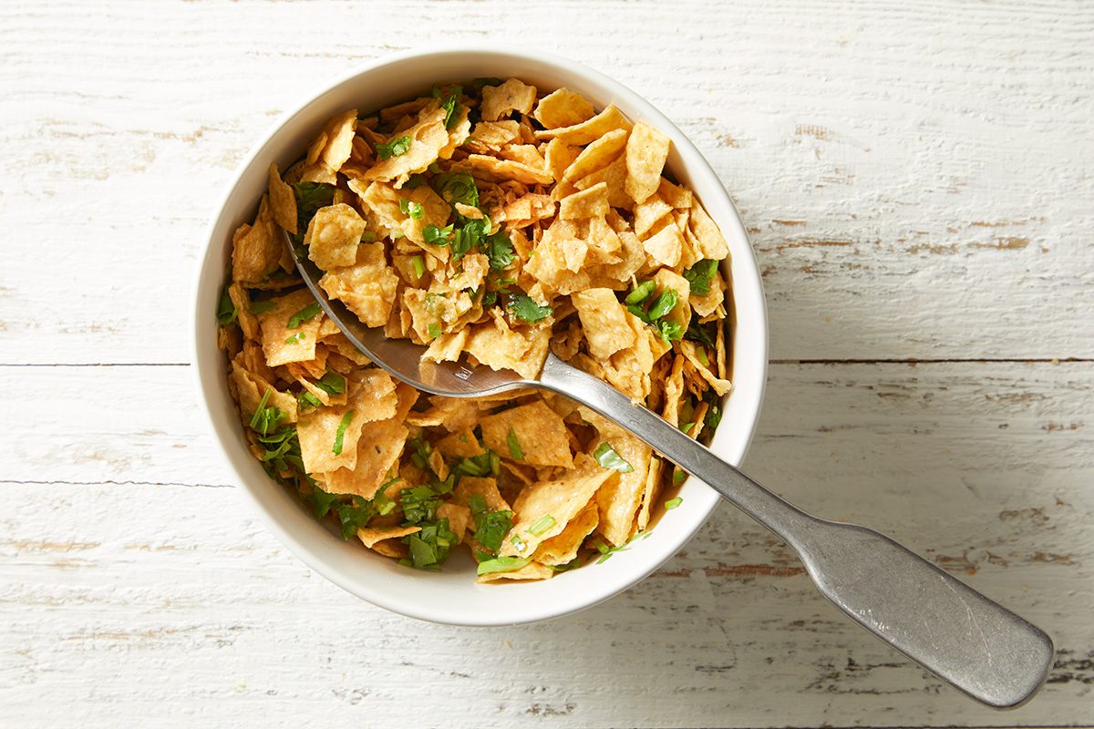 A white bowl filled with crushed crackers or chips topped with chopped green herbs, with a spoon resting inside, placed on a light wooden surface.