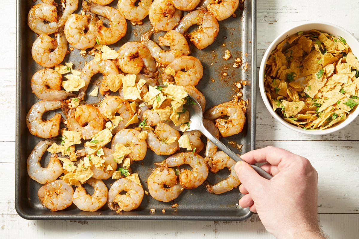 A hand sprinkles crushed tortilla chips over raw seasoned shrimp arranged on a baking sheet, with a bowl of crushed chips nearby on a white wooden surface.