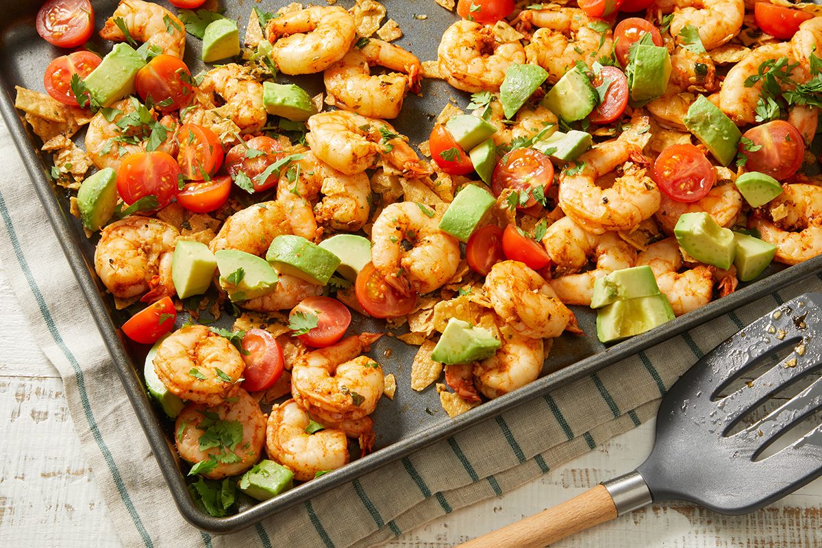 A sheet pan filled with cooked shrimp, diced avocado, cherry tomato halves, herbs, and tortilla chips sits on a striped kitchen towel next to a serving spatula.