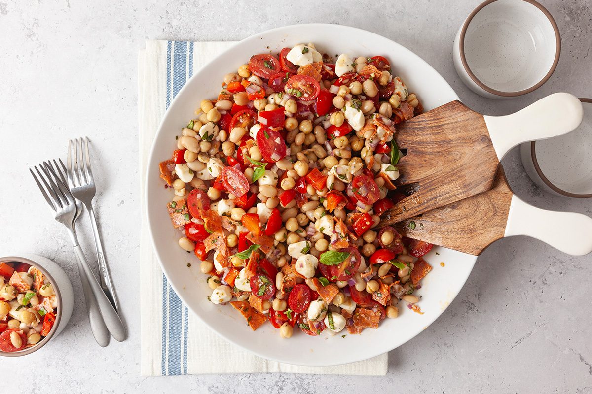 A large white bowl filled with a colorful salad of chickpeas, cherry tomatoes, mozzarella balls, herbs, and chopped vegetables sits on a striped napkin with salad servers, forks, and small bowls nearby.