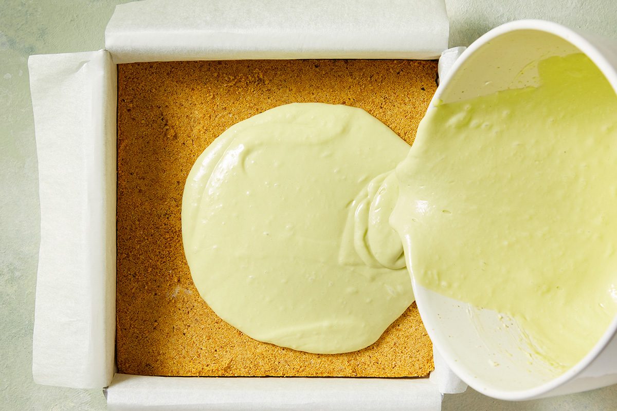 A bowl is pouring pale green batter onto a square pan lined with parchment paper, covering a layer of crumbly crust.