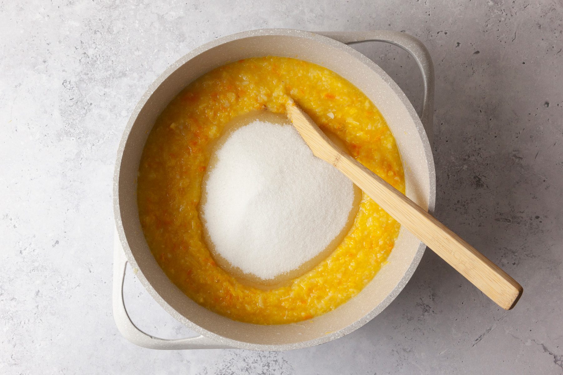 overhead shot of a pot filled with a yellowish mixture that appears to be a blend of ingredients such as squash; in the center of the mixture, there is a mound of white sugar; a wooden spatula rests against the edge of the pot, the background features a subtle gray surface