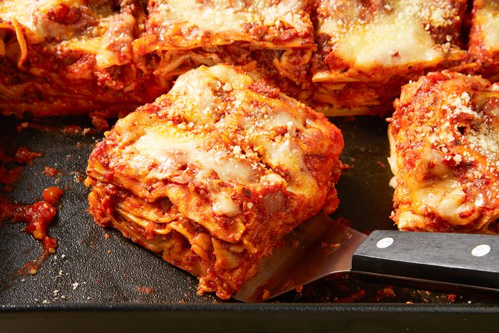 A close-up of a serving of lasagna on a spatula resting in a baking dish. The lasagna has layers of pasta, rich tomato sauce with meat, and melted cheese, topped with grated parmesan. Some slices remain in the dish.