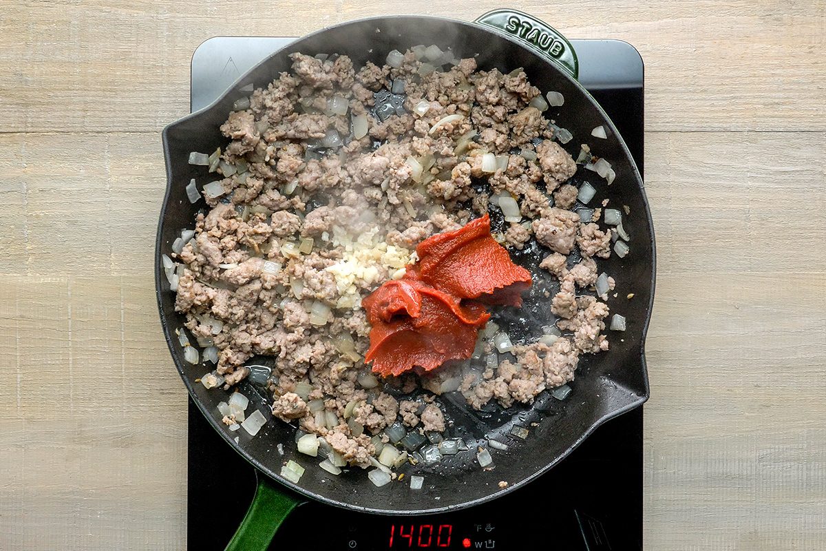 A frying pan on an induction stove contains ground meat, diced onions, garlic, and two dollops of tomato paste. Steam rises from the mixture as it cooks. The stove display shows "1400" indicating the heat setting or time.