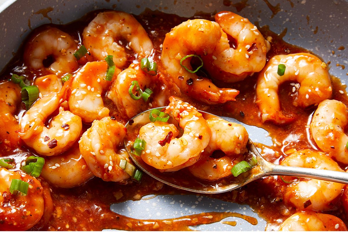 Shrimp cooked in a spicy, reddish sauce, garnished with chopped green onions, with a spoon resting in the pan.