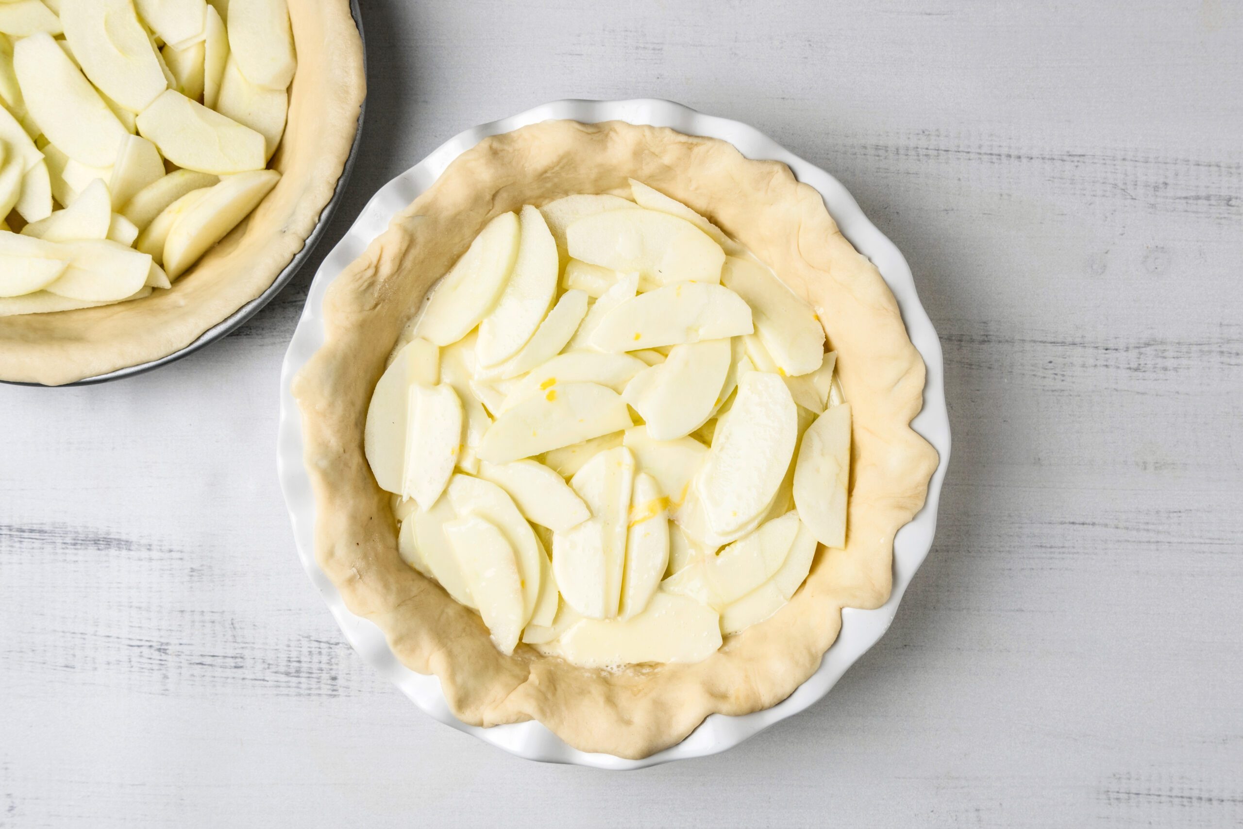 Unbaked pie with sliced apples arranged in a crust placed in a white pie dish on a light gray surface. Another pie is partially visible in the background.