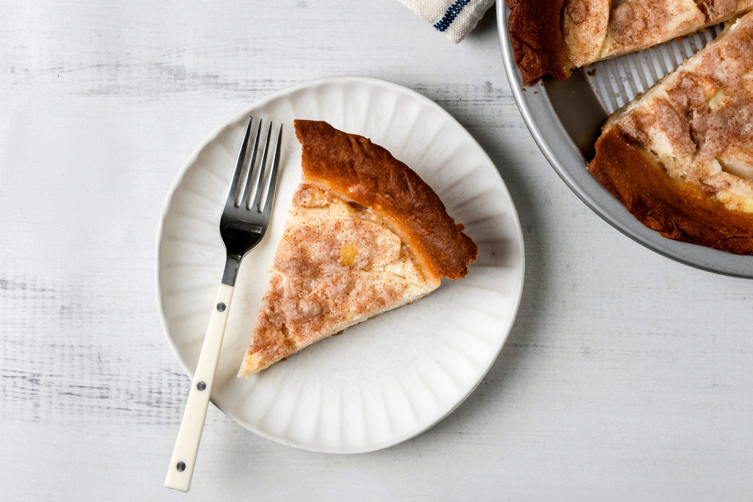 A slice of apple pie on a white plate with a fork beside it. The pie has a golden-brown crust with visible cinnamon sugar on top. The rest of the pie is in a silver baking dish in the background on a white surface.