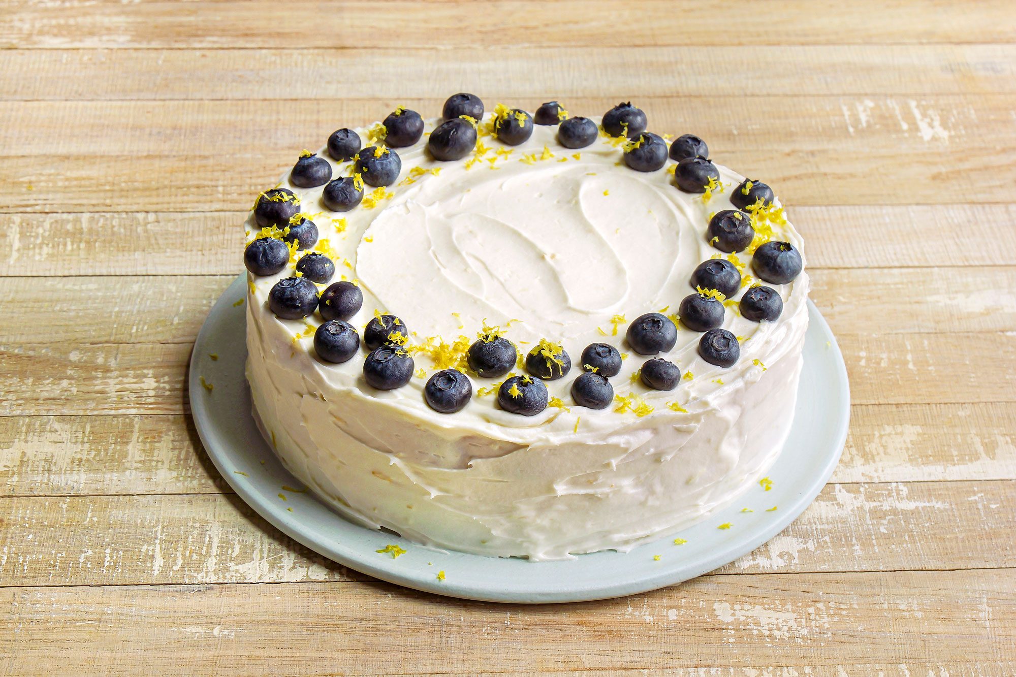 3/4th shot of a lemon blueberry cake displayed on a pale plate, the top is garnished with fresh blueberries arranged in a circular pattern, and lemon zest sprinkled around the berries, the cake rests on a rustic wooden surface