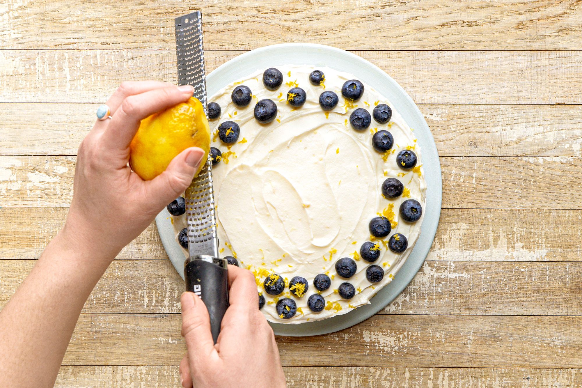 overhead shot of a hand is shown zesting a lemon over a cake that is decorated with a circular arrangement of blueberries and lemon zest; the surface beneath the cake is made of light colored wooden planks