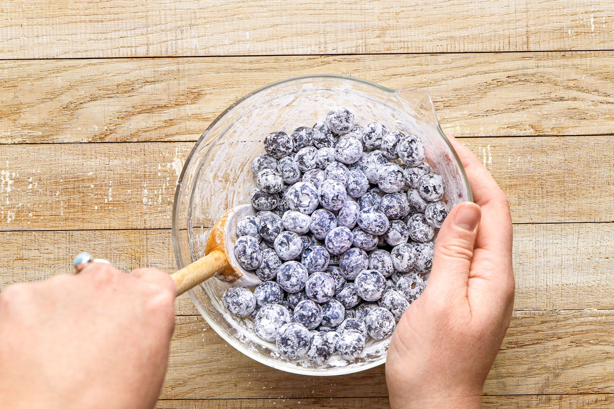 overhead shot of a person’s hands mixing a bowl of blueberries coated in a white powdery substance or sugar, the bowl is transparent and made of glass, the background consists of wooden texture top