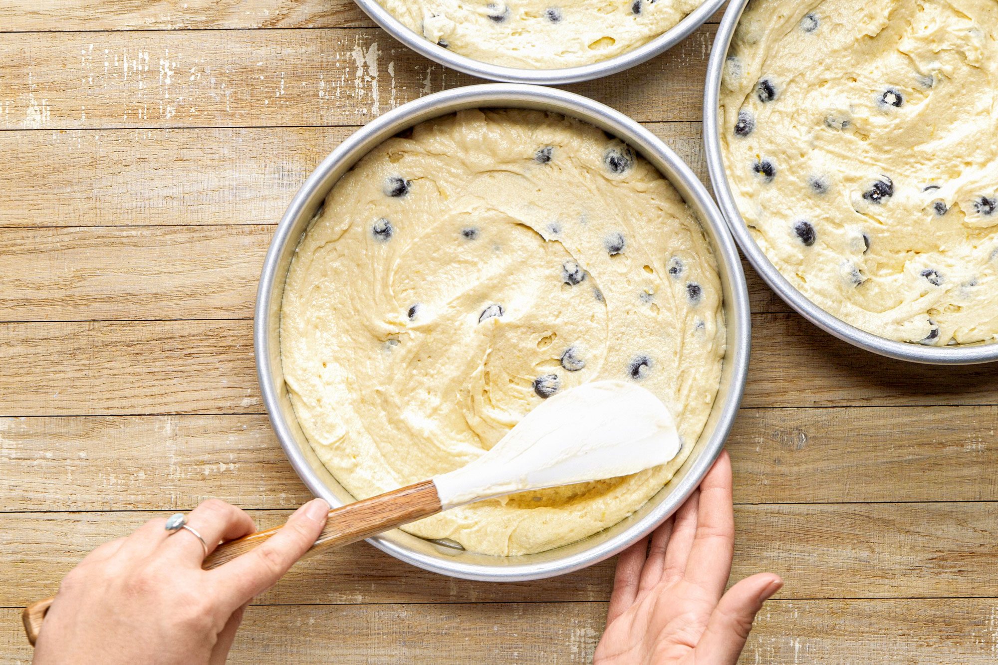 overhead shot of three round cake pans filled with a light colored batter that is dotted with black blueberries; one of the pans is being adjusted with a spatula by a hand that is partially visible in the frame; the background is wooden surface