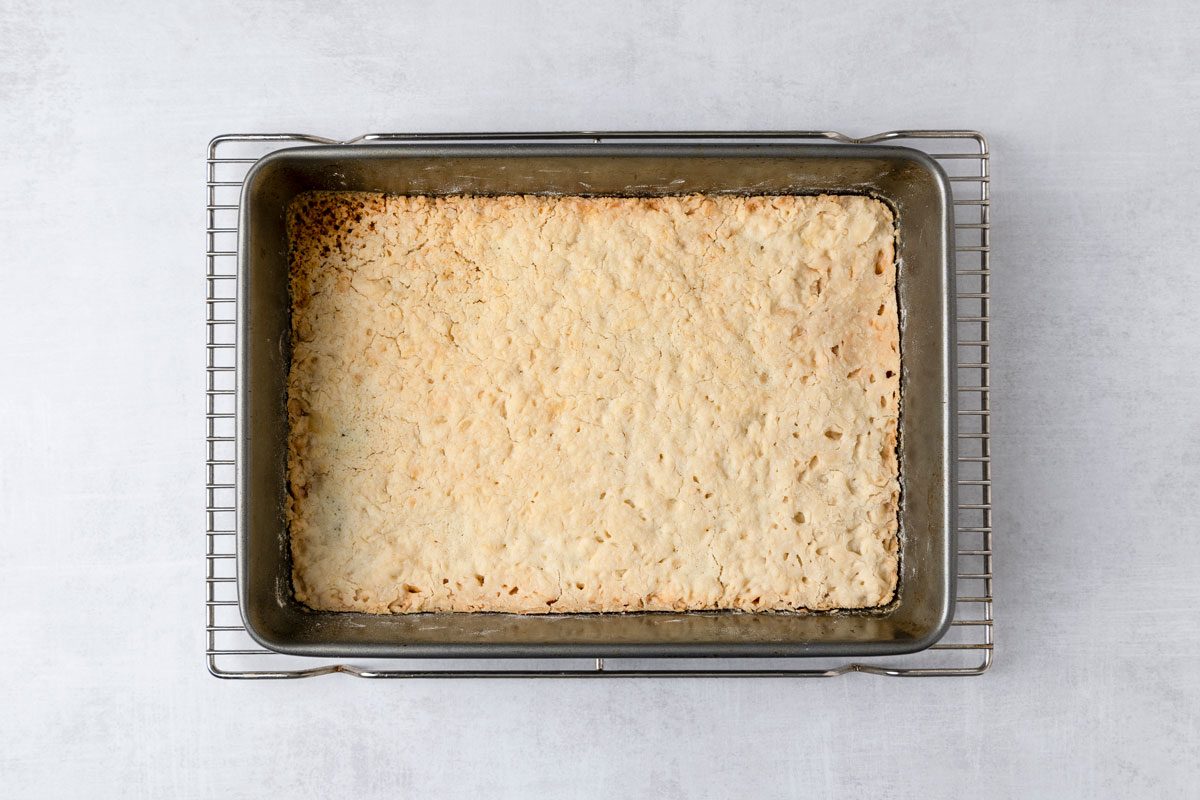 overhead shot of a baking pan placed on a cooling rack; the pan contains a large slab of baked goods, the pan itself is metallic and rectangular, while the surface beneath is smooth and light colored