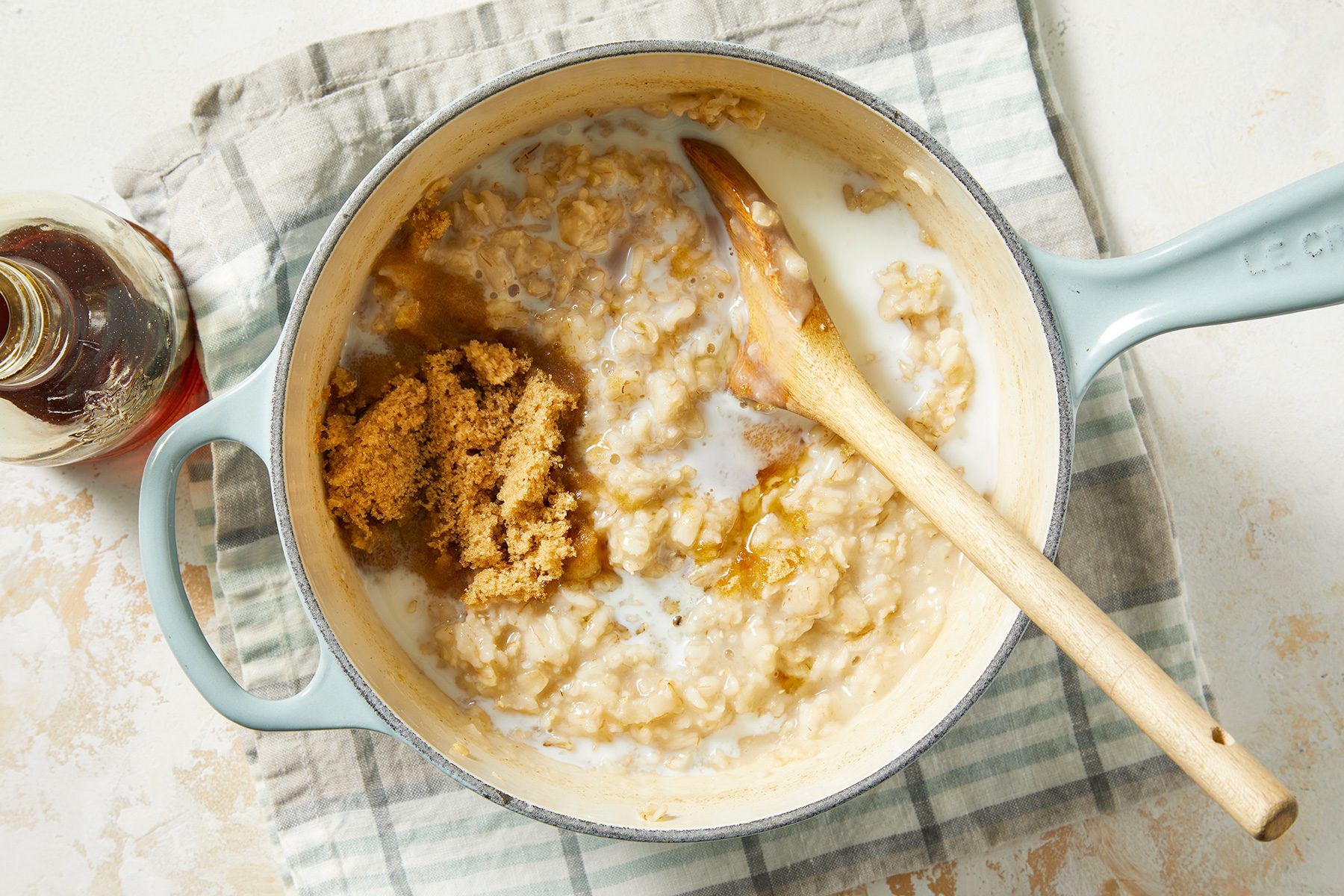 A blue pot filled with cooked oatmeal, topped with brown sugar and a wooden spoon, rests on a checkered cloth. A small glass jar of syrup is nearby on a light textured surface.