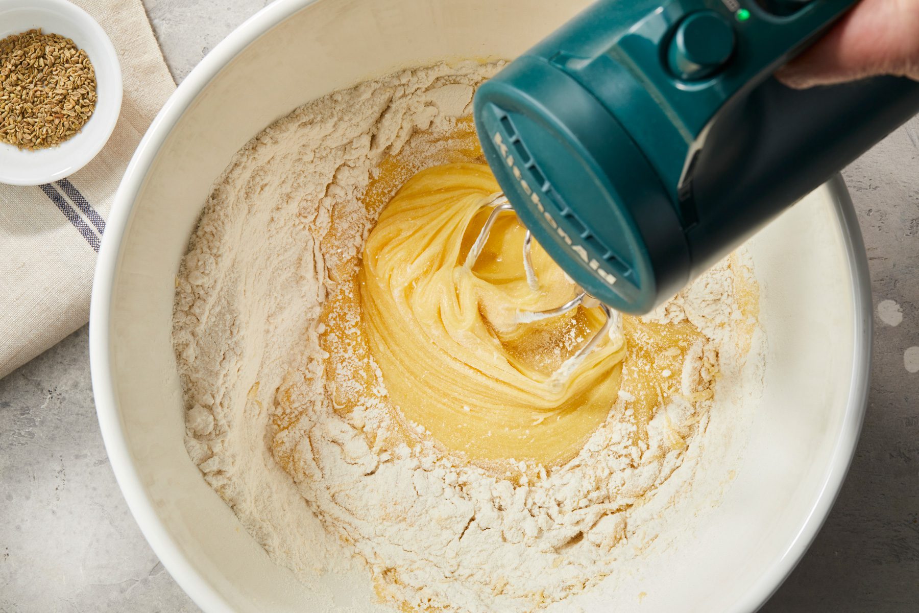 overhead shot of a mixer is blending a creamy yellow batter inside a large white mixing bowl; there is a small white dish containing what appears to be fennel seeds; the countertop surface is gray