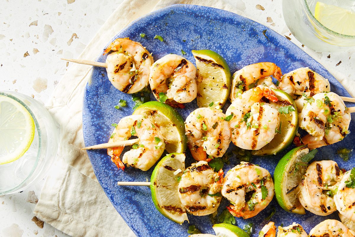 Grilled shrimp skewers with lime slices arranged on a blue plate. The shrimp are seasoned and garnished with herbs. Two glasses with lemon slices are visible in the background on a light-colored surface.