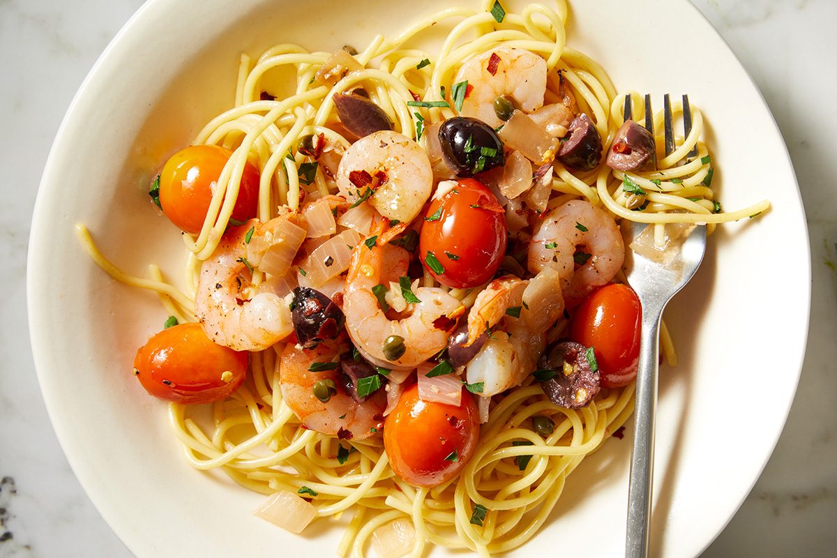 A plate of spaghetti topped with shrimp, cherry tomatoes, black olives, and fresh herbs. A fork rests on the side of the dish.
