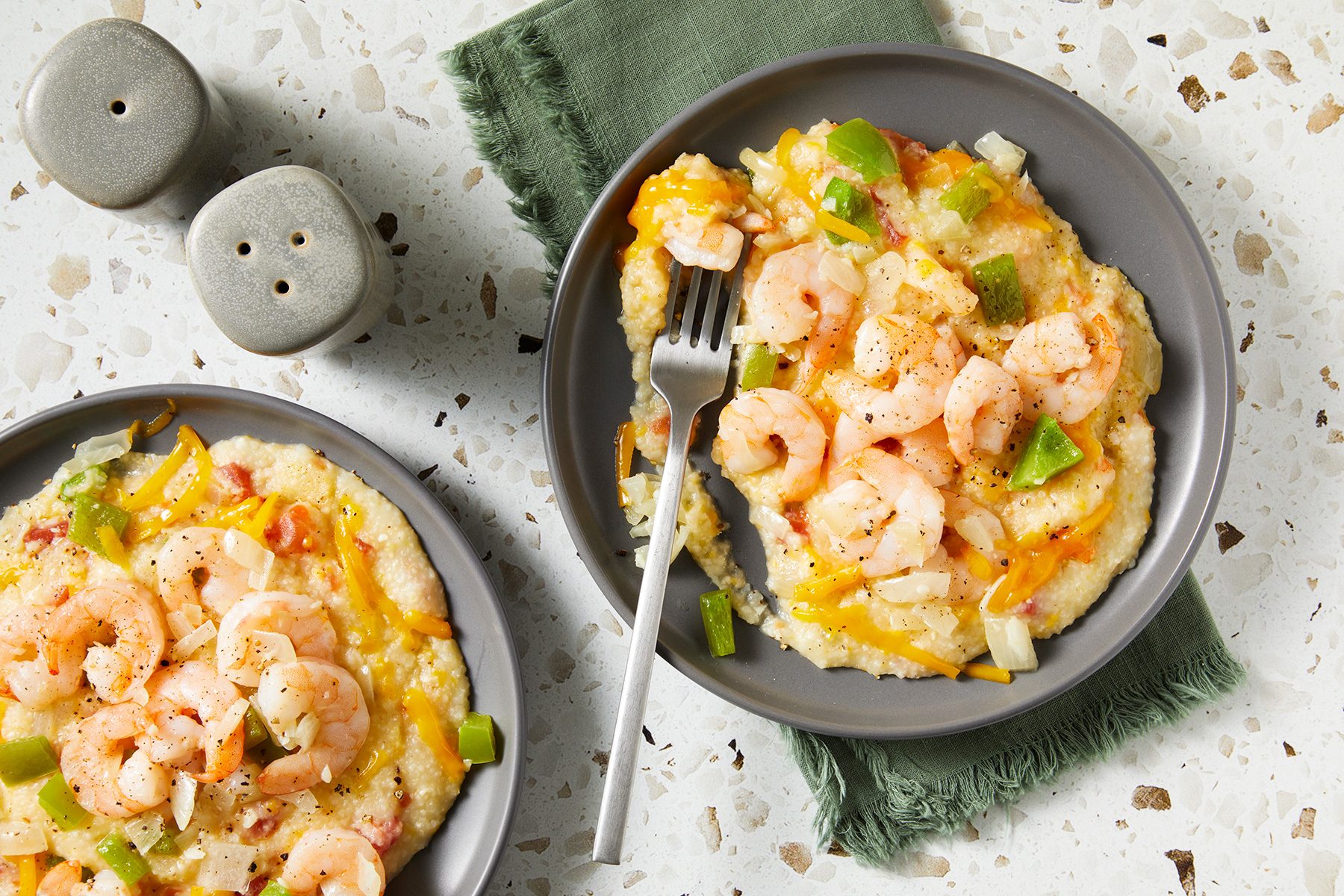 Plates of shrimp and grits topped with green peppers, onions, and melted cheese. A fork rests on one plate. 