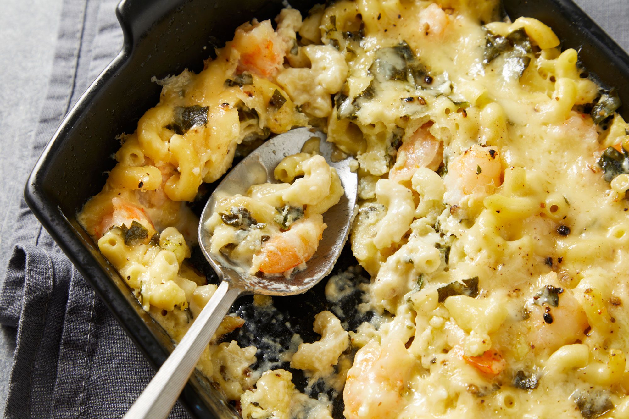 Close shot of Shrimp & Macaroni Casserole; in a black baking pan placed over grey napkin; with a serving spoon; grey surface;