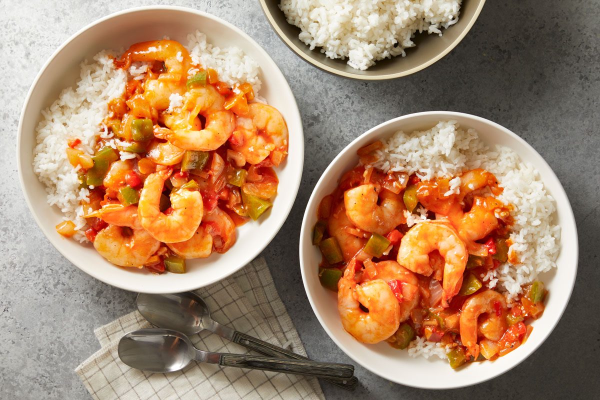 overhead shot of two white bowls of Spicy Shrimp with Rice; in the foreground, two silver spoons lay on a beige checkered napkin, in the background, a third bowl of rice is visible