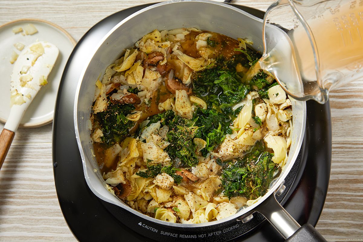 A pot on a stove contains a mixture of noodles, leafy greens, mushrooms, and chicken pieces. A hand is pouring broth from a pitcher into the pot, and a spatula with some garlic is resting on a small plate nearby.