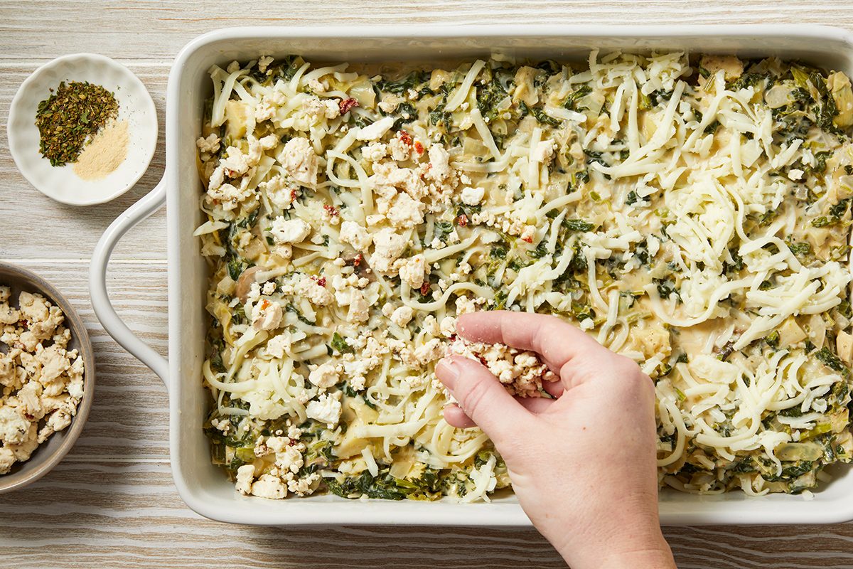A hand is sprinkling breadcrumbs over a casserole in a rectangular baking dish. The casserole is topped with shredded cheese and appears to contain various ingredients like vegetables. A bowl of spices and a bowl of breadcrumbs are nearby.