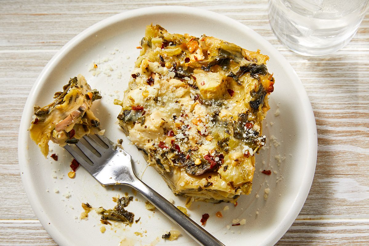 A white plate holds a square piece of lasagna with visible layers of pasta, spinach, cheese, and sauce. A fork rests beside it, and a glass of water is partially visible in the top right corner. The table underneath is light wood.