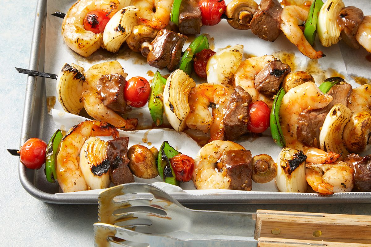 Grilled skewers with shrimp, beef, onions, cherry tomatoes, and green bell peppers on a baking tray, with metal tongs placed nearby.