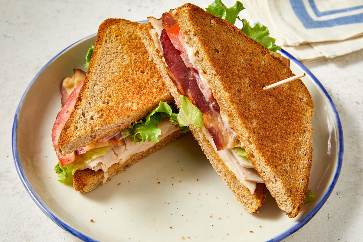 A plate with a toasted turkey club sandwich cut in half, showing layers of lettuce, tomato, bacon, turkey, and bread. One half is held together with a toothpick. 