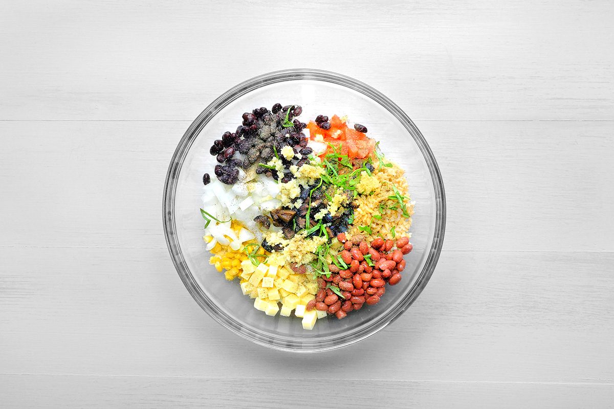 overhead shot of a glass bowl contains a vibrant and colorful mixture of ingredients for a Mexican Street Corn Salad, Black beans, corn kernels, diced red onion, chopped cilantro, shredded cheddar cheese, and kidney beans are combined in the bowl