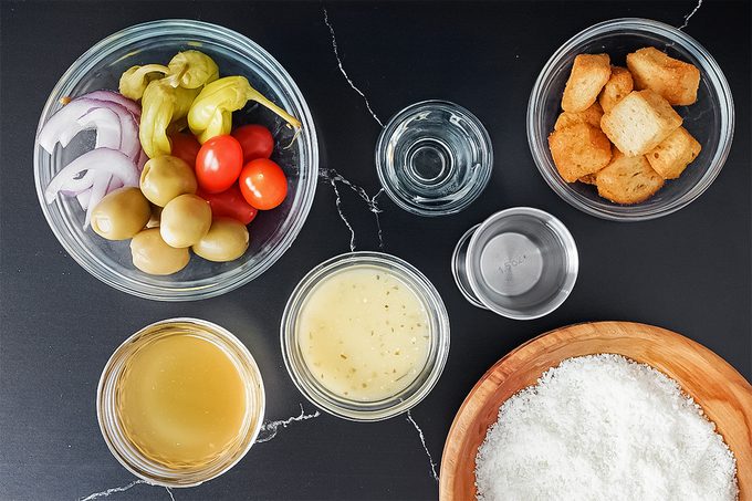 ViralaVarious ingredients in bowls on a dark surface, including sliced onions, olives, cherry tomatoes, dressing, croutons, and grated cheese. Two smaller bowls contain liquids.Olive Garden Salad Martini Lauren Habermehl For Taste Of Home Trends 02 Yvedit A