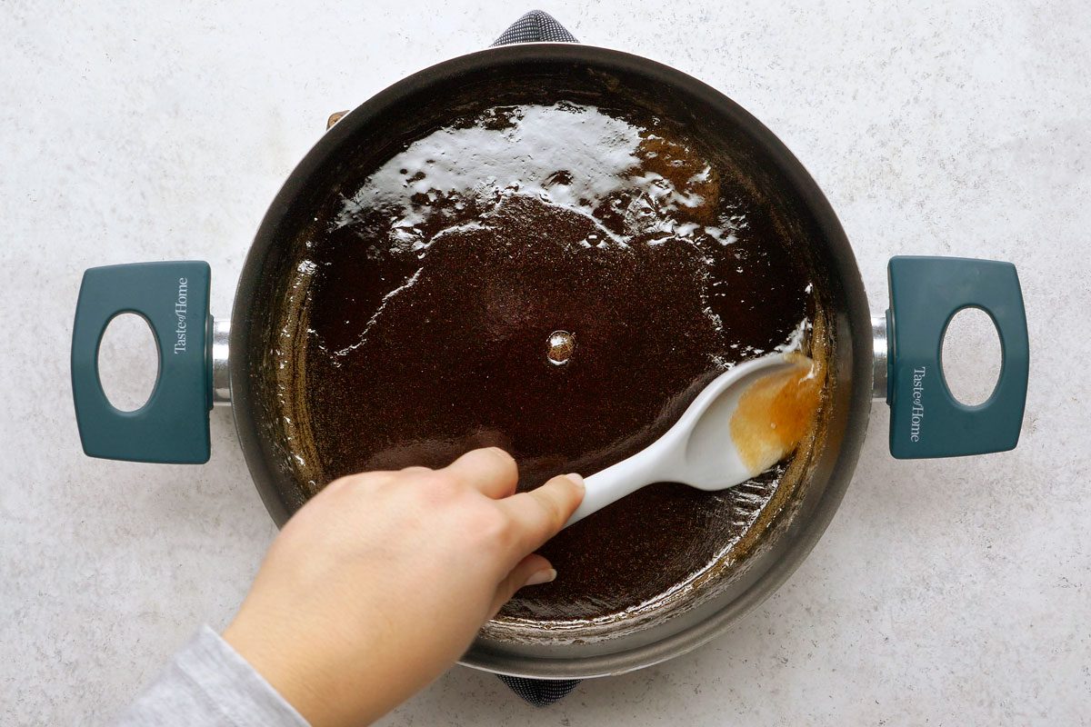 overhead shot of a hand stirs a dark brown liquid in a metal pan with a white spatula; the pan is surrounded by a white surface and has a green handle on each side; the liquid has a shiny and glossy appearance, giving the impression that it is a melted mixture or a caramel sauce;