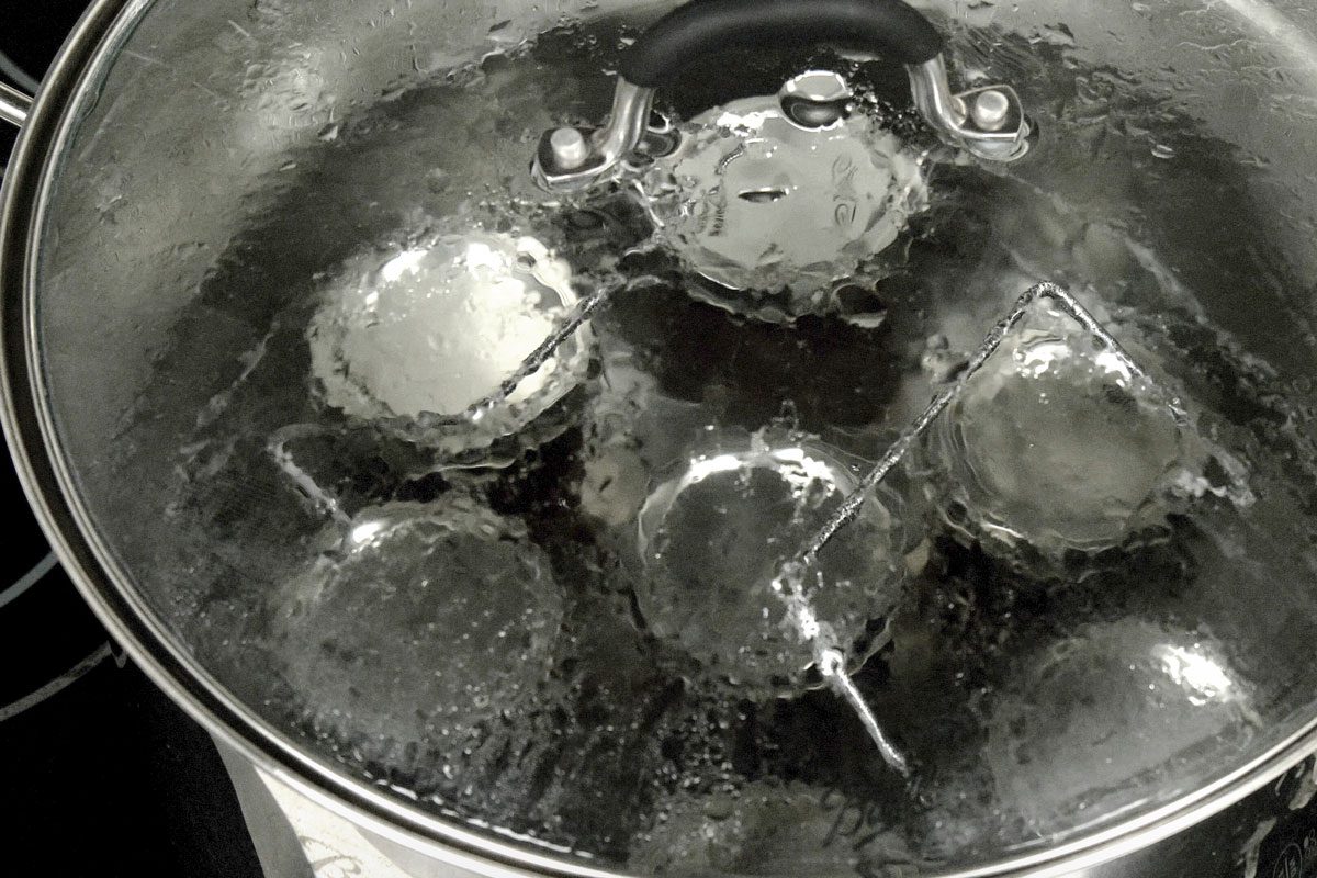 3/4th shot of a pot with four glass jars inside is on a stovetop; the pot is filled with boiling water; the water is bubbling and churning inside the pot; the jars are submerged in the water and the pot is covered with a lid; the lid is secured to the pot by a handle that has a large knob on top; there is a metal piece that is connected to the lid by a metal bar; the metal piece is also secured to the handle; the metal piece is used to hold the jars in place inside the pot; the whole pot is made of stainless steel and has a shiny finish;