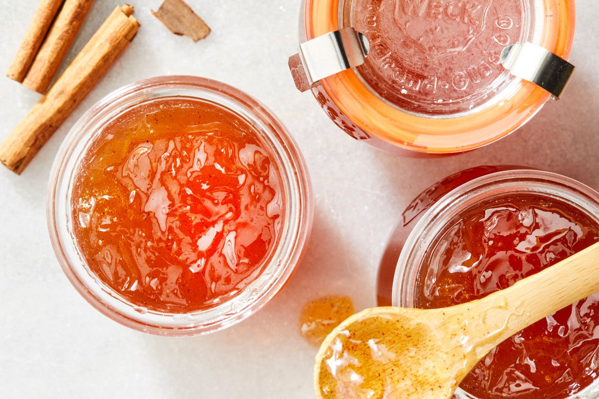 Top view shot of Apple Cinnamon Jelly; stored in glass jars; with a spoon resting over one jar; and cinnamon sticks scattered in the background; all arranged on a marble surface;